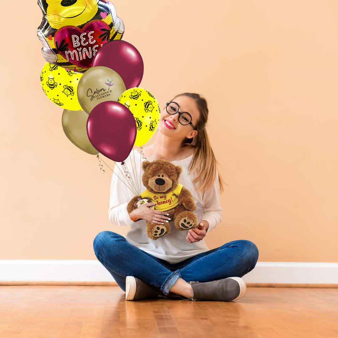 A girl sitting on the floor as she holds a Valentine's balloon bouquet with teddy bear holding a bee mylar balloon and a set of latex helium balloons. His T-shirt has a 