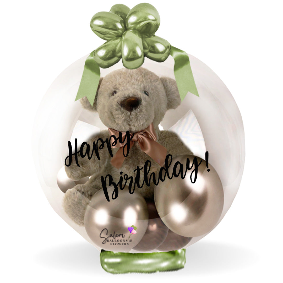 Beige teddy bear in a clear bubble balloon decorated with ribbon and a "Happy Birthday" message. Salem Oregon balloon delivery.