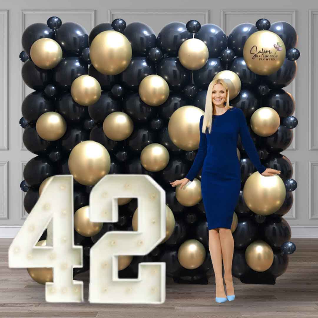A woman posing for a picture in front of a black and gold Balloon wall with light up marquee numbers. Salem Oregon balloon decoration.