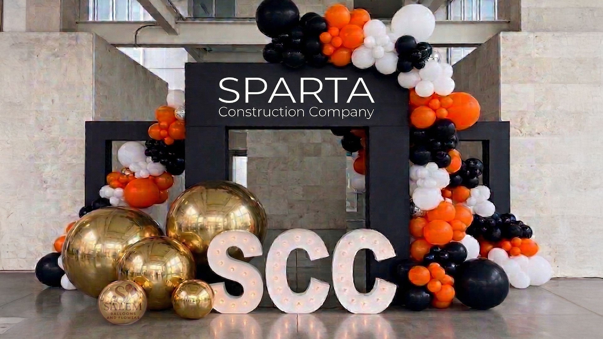 Large balloon decor set with 3 large black square arches decorated with large balloon garlands in black, orange, and white with giant gold spheres and 3 marquee letters spelling SCC with soft yellow lights. Corporate event decor. Oregon.
