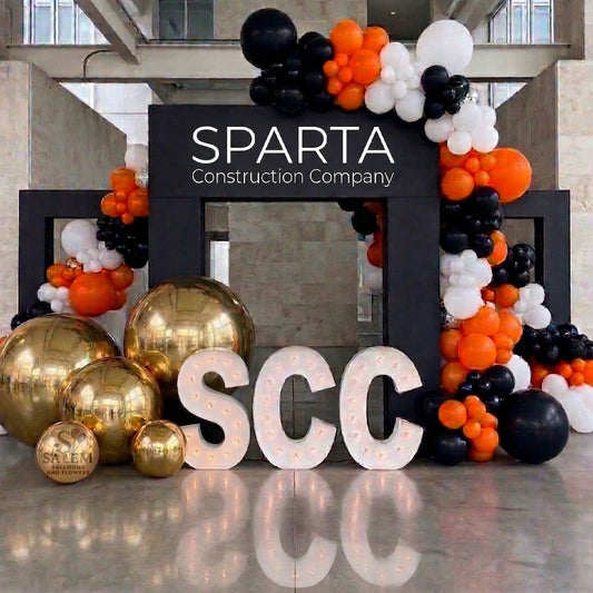 Large balloon decor set with 3 large black square arches decorated with large balloon garlands in black, orange, and white with giant gold spheres and 3 marquee letters spelling SCC with soft yellow lights. Corporate event decor. Oregon.