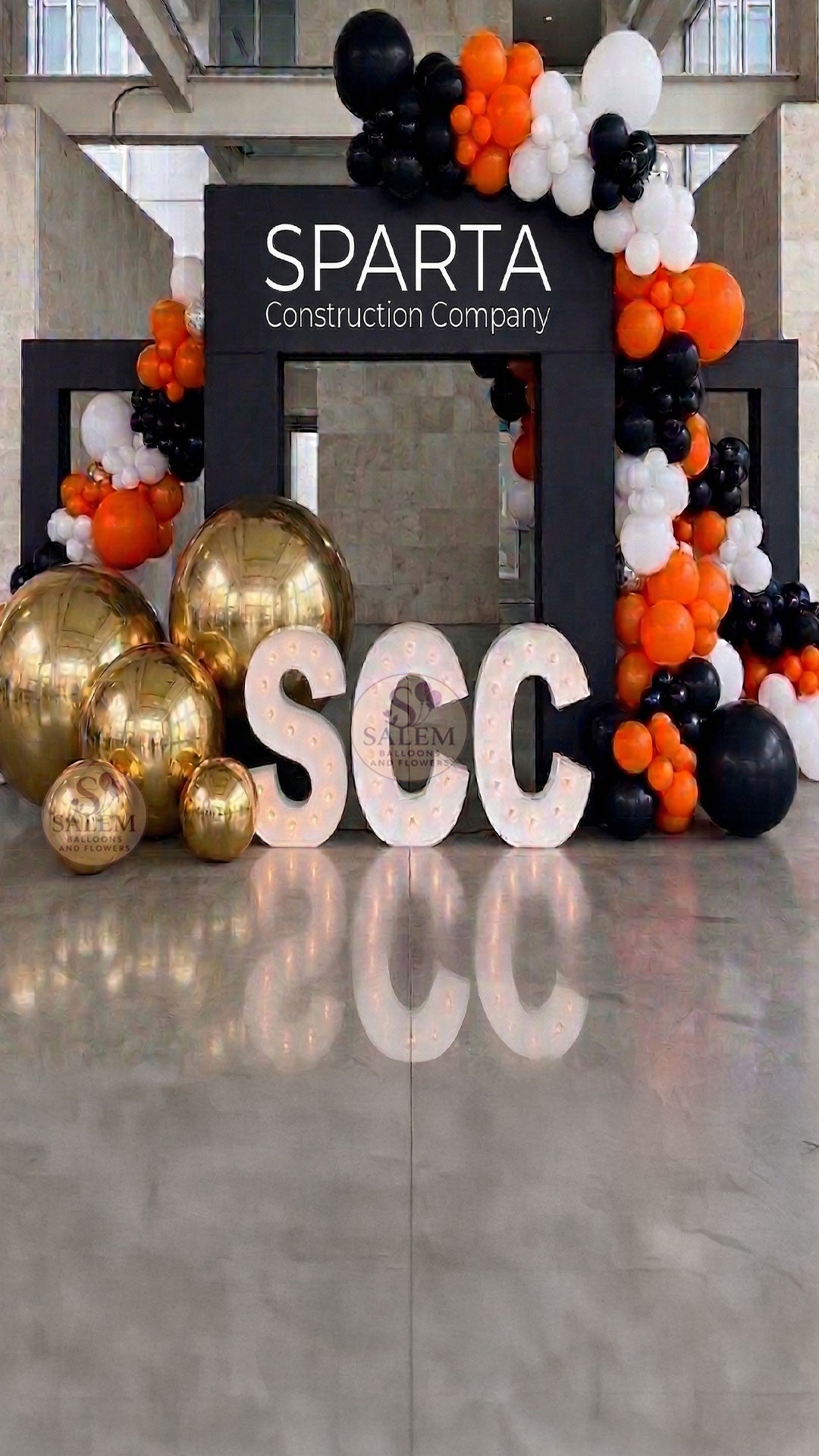 Large balloon decor set with 3 large black square arches decorated with large balloon garlands in black, orange, and white with giant gold spheres and 3 marquee letters spelling SCC with soft yellow lights. Corporate event decor. Oregon.