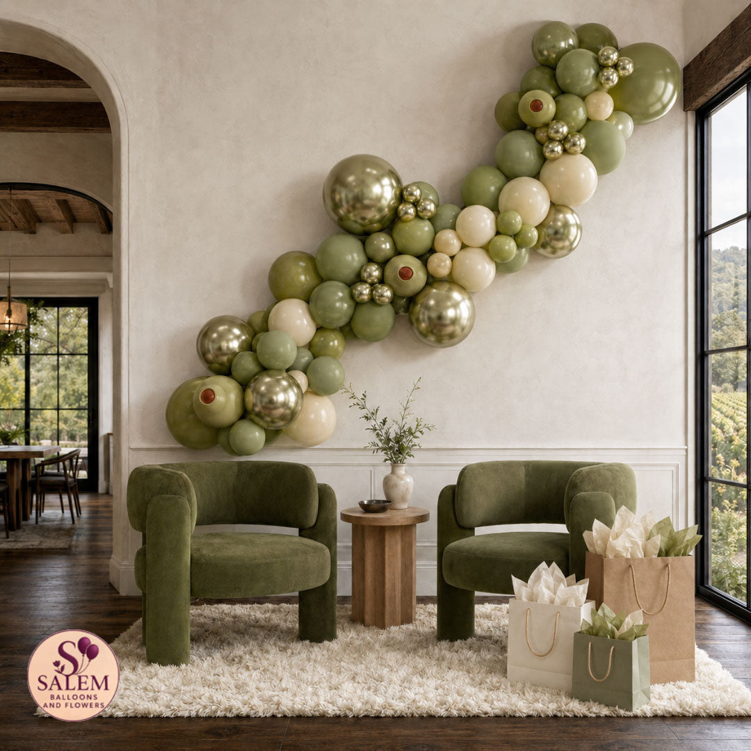 Olive-green, sage, cream, and metallic specialty balloon garland above two green lounge chairs in a neutral vineyard-style home. With a Salem Balloons and Flowers logo on the left bottom corner.