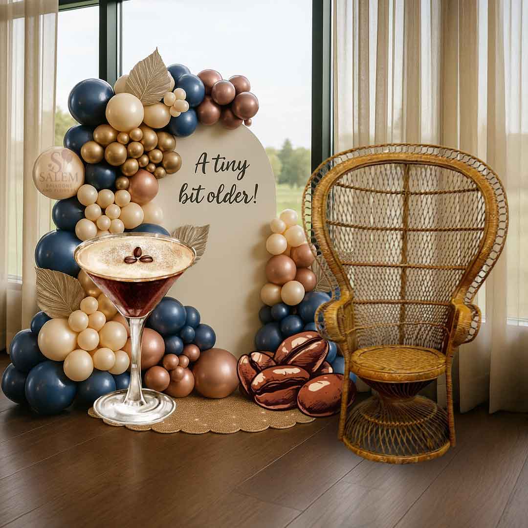2 balloon garland in navy blue, beige and mocha, decorated with cream palm leaves decorating a beige arch panel with a "A tiny bit older" sign and 2 large espresso Martini and coffee beans cutouts, next to a wicker picklock chair. Balloon decor Oregon. 