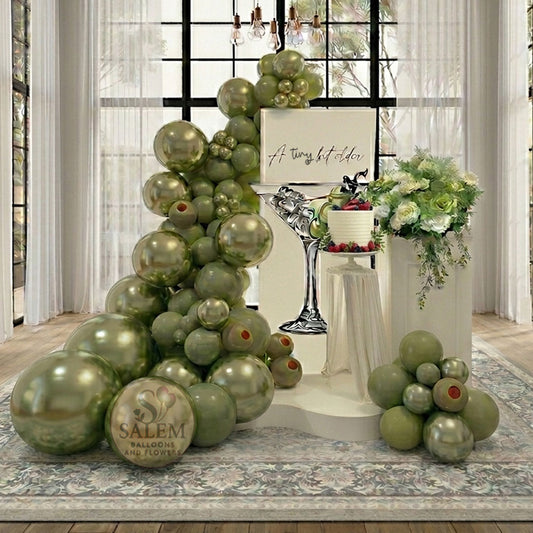 A tiny bit older themed balloon decor. Featuring a large balloon garland in olive green with a square ivory panel with a martini cup cutout and an "A tiny bit older" message, decorated with flowers on a pedestal  in a loft with tall windows. Balloon decor. Oregon.