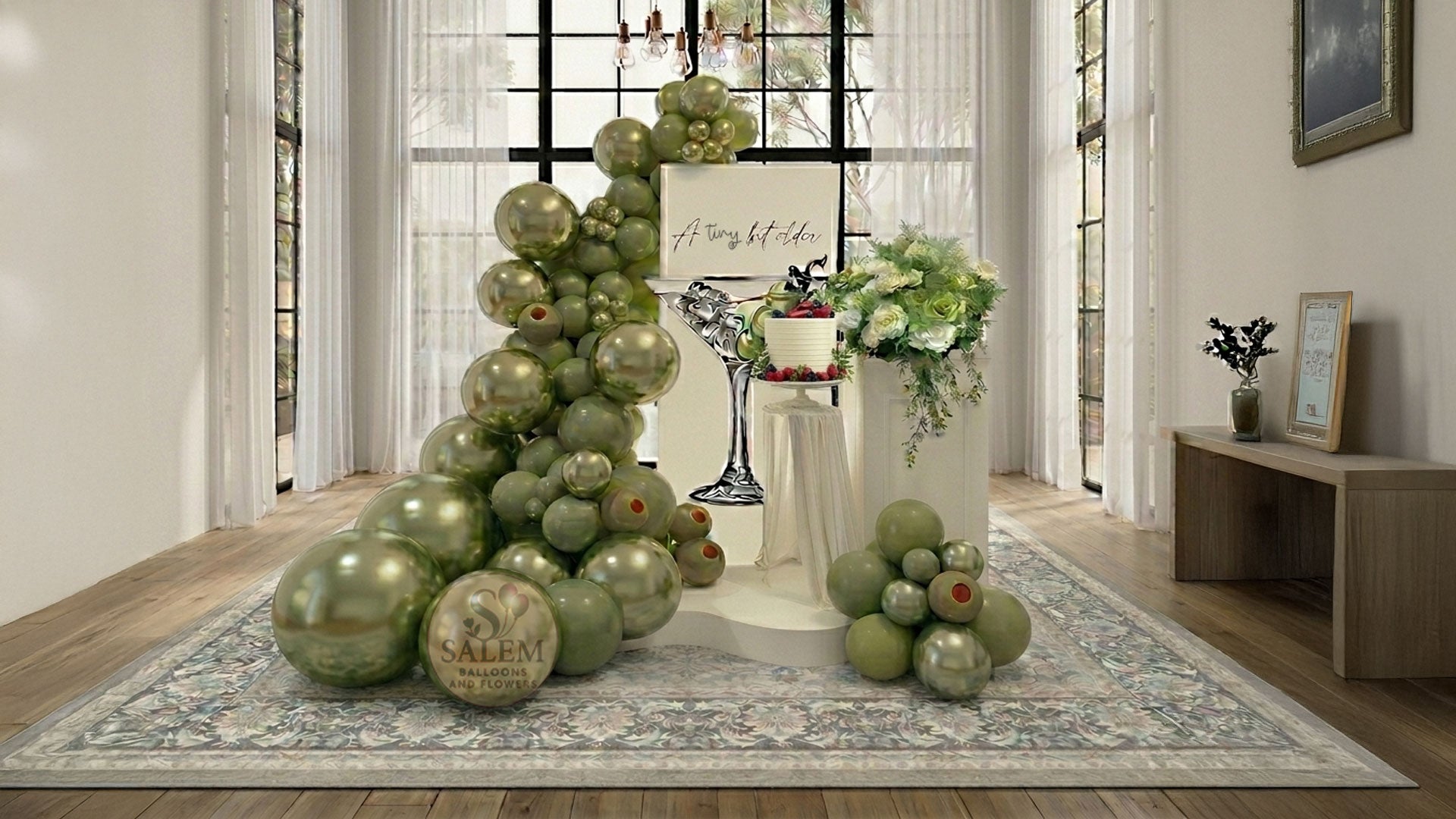 A tiny bit older themed balloon decor. Featuring a large balloon garland in olive green with a square ivory panel with a martini cup cutout and an "A tiny bit older" message, decorated with flowers on a pedestal  in a loft with tall windows. Balloon decor. Oregon.