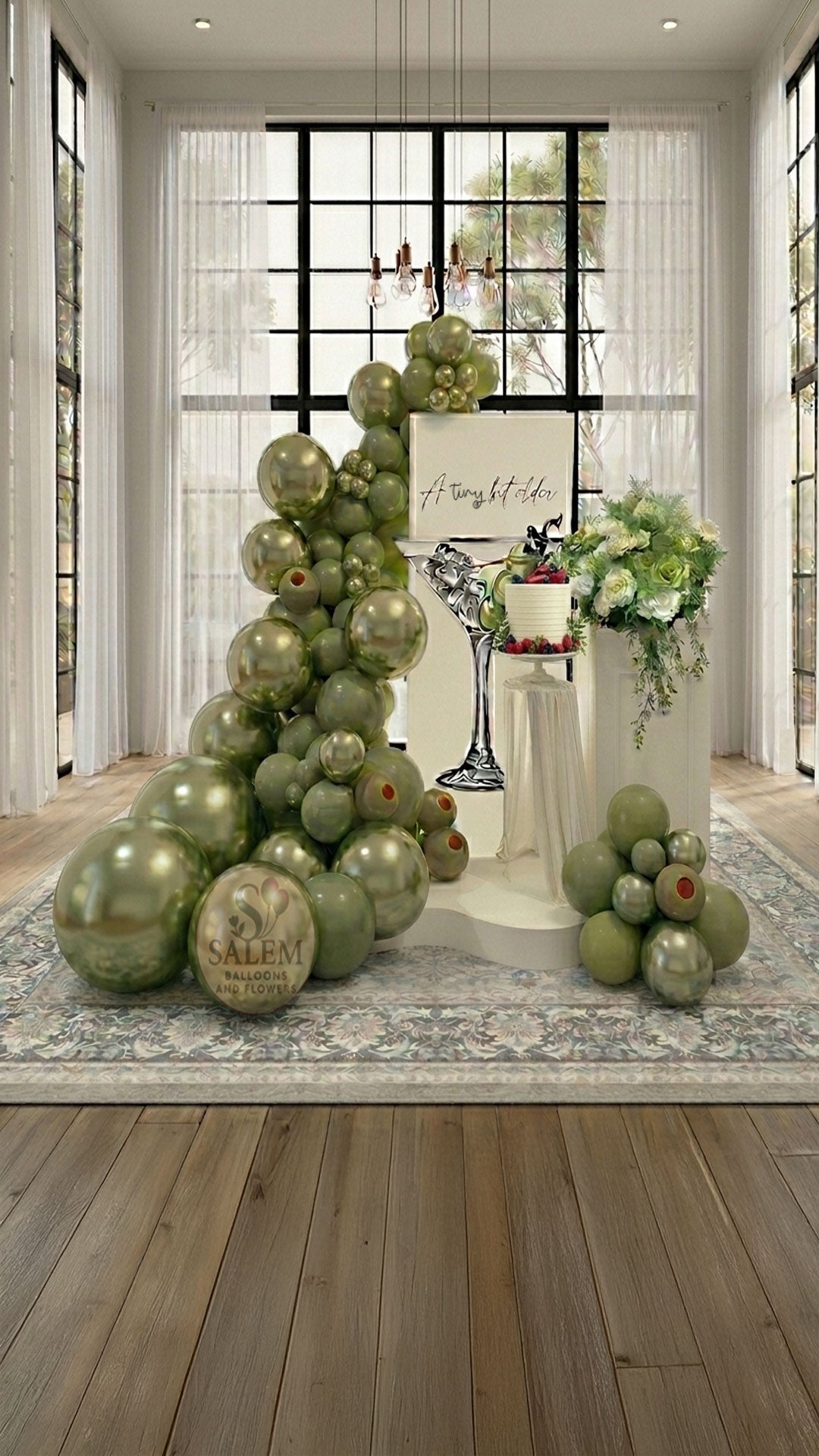 A tiny bit older themed balloon decor. Featuring a large balloon garland in olive green with a square ivory panel with a martini cup cutout and an "A tiny bit older" message, decorated with flowers on a pedestal  in a loft with tall windows. Balloon decor. Oregon.