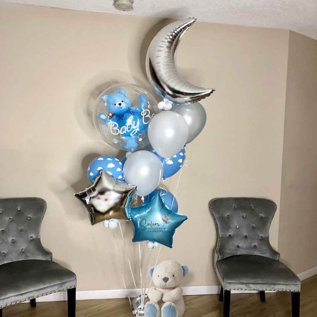 An "It's a Boy" Balloon bouquet in silver, pastel blue and white. Featuring a stuffed bubble balloon with a blue teddy bear, a large moon and stars standing in a living room. Balloon delivery in Salem Oregon and nearby cities.