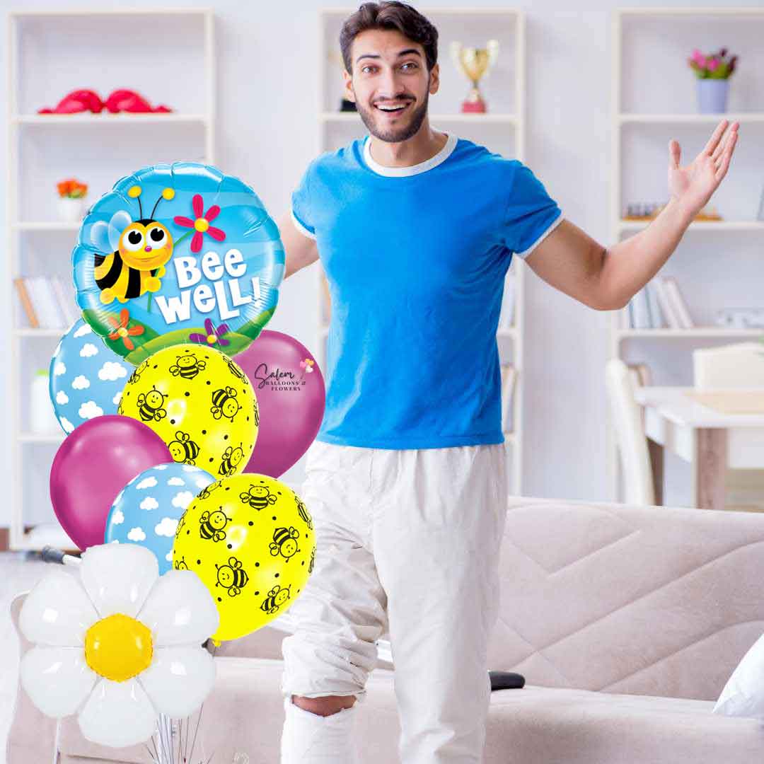 A guy recovering at home standing next to Get well helium balloon bouquet, featuring a mylar balloon with a cute bee and a 'Bee Well!