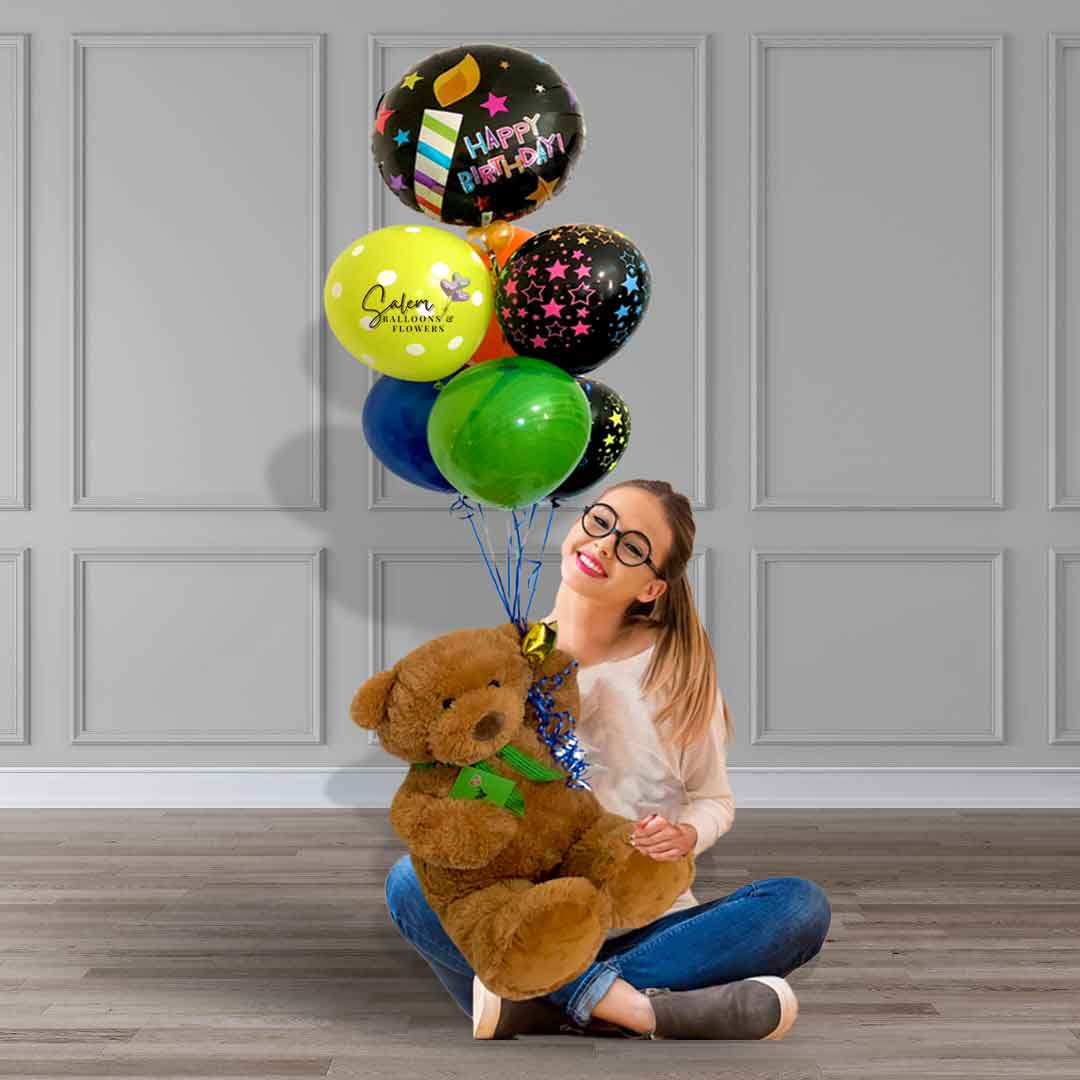 A young girl sitting on the floor holding a Large teddy bear holding a set of helium-filled balloons with a 