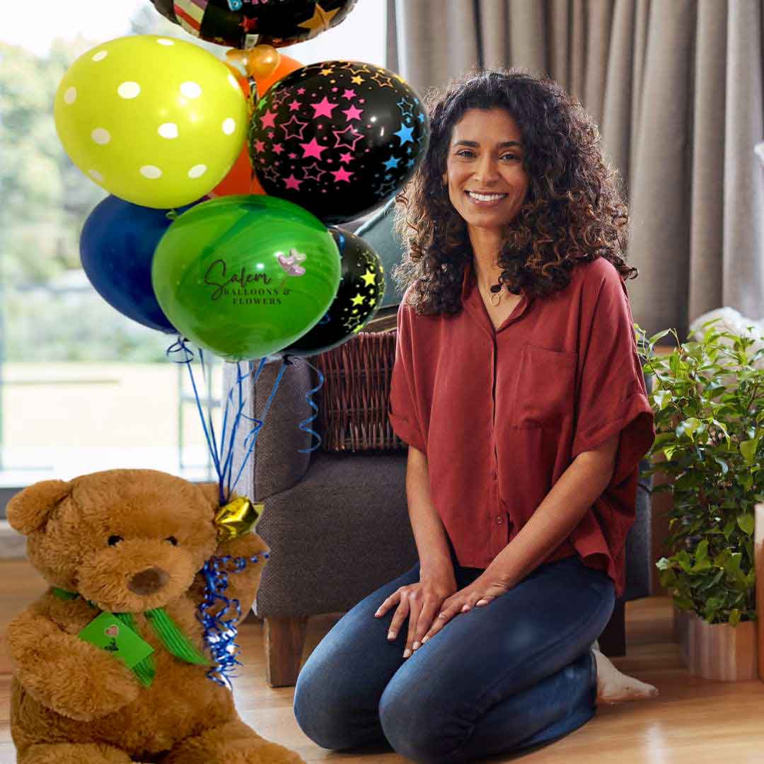A woman kneeling on the floor next to a Large teddy bear holding a set of helium-filled balloons with a "Happy Birthday" message. Balloon delivery in Salem Oregon and nearby cities.