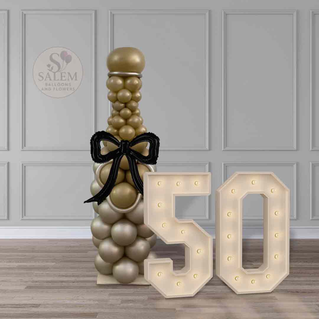 Champagne-inspired balloon column with gold balloons and black bow, standing in front of a gray wall with Marquee numbers #50. Luxury balloon decor by Salem Balloons and Flowers in Oregon.