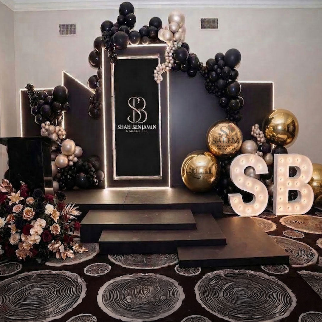 Corporate decor setup with balloon garlands, flowers, and 'SB' letter lights, 5 lit black panels on 4 black platform in different heights in a venue room. Event decor Oregon.