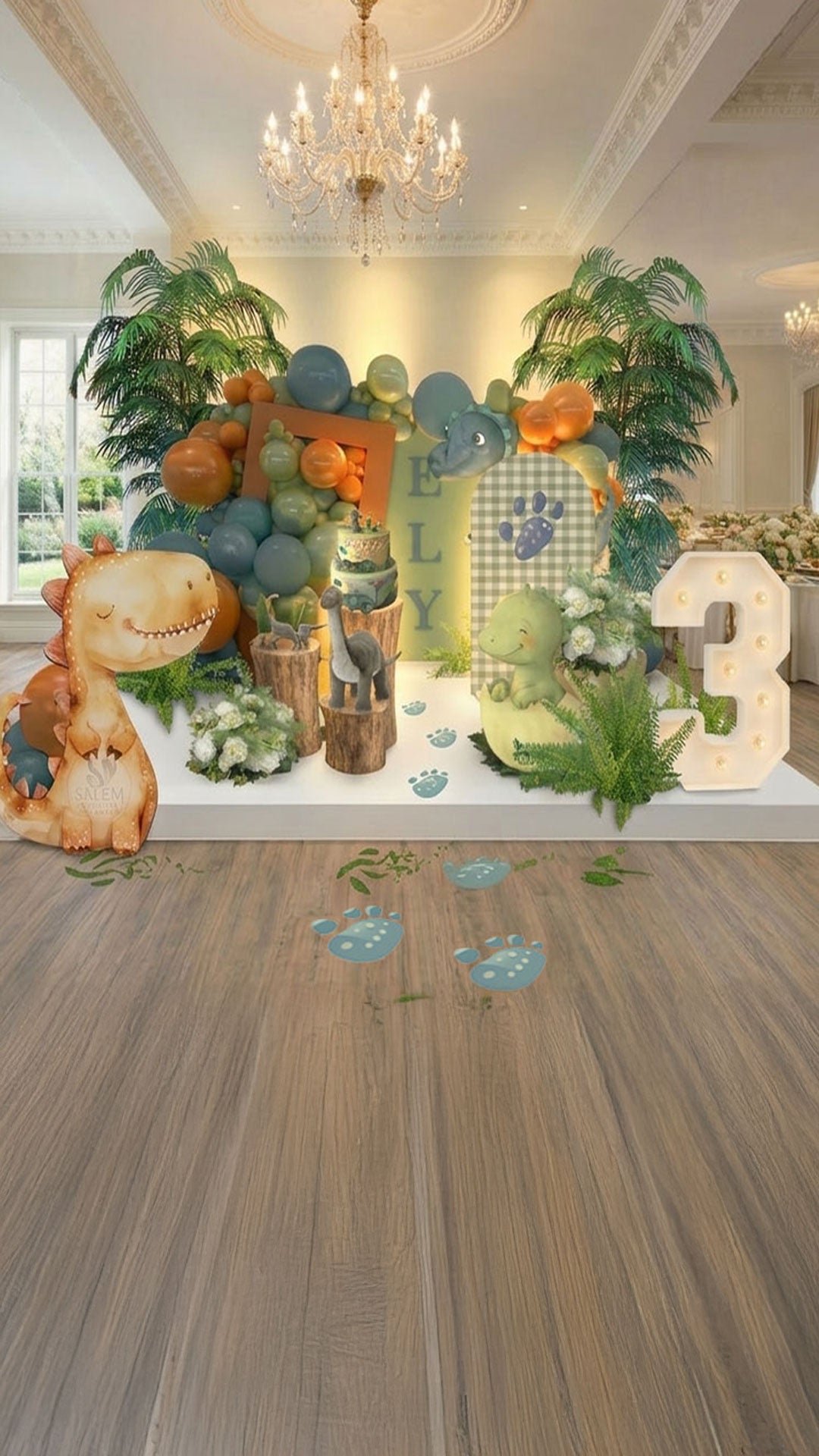 Dinosaur-themed birthday party setup with decorations in a green blue and orange color palette, decorated with greenery, palms, dinosaurs cutouts, marquee number 3 in an elegant venue room with chandeliers. Balloon Decor Oregon.