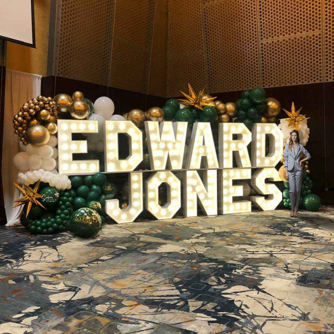 Light-Up marquee letters display spelling Edward Jones, nested in a 10 ft tall organic balloon wall in gold, green, and white. Salem Oregon Balloon Decor at the Salem Convention Center.