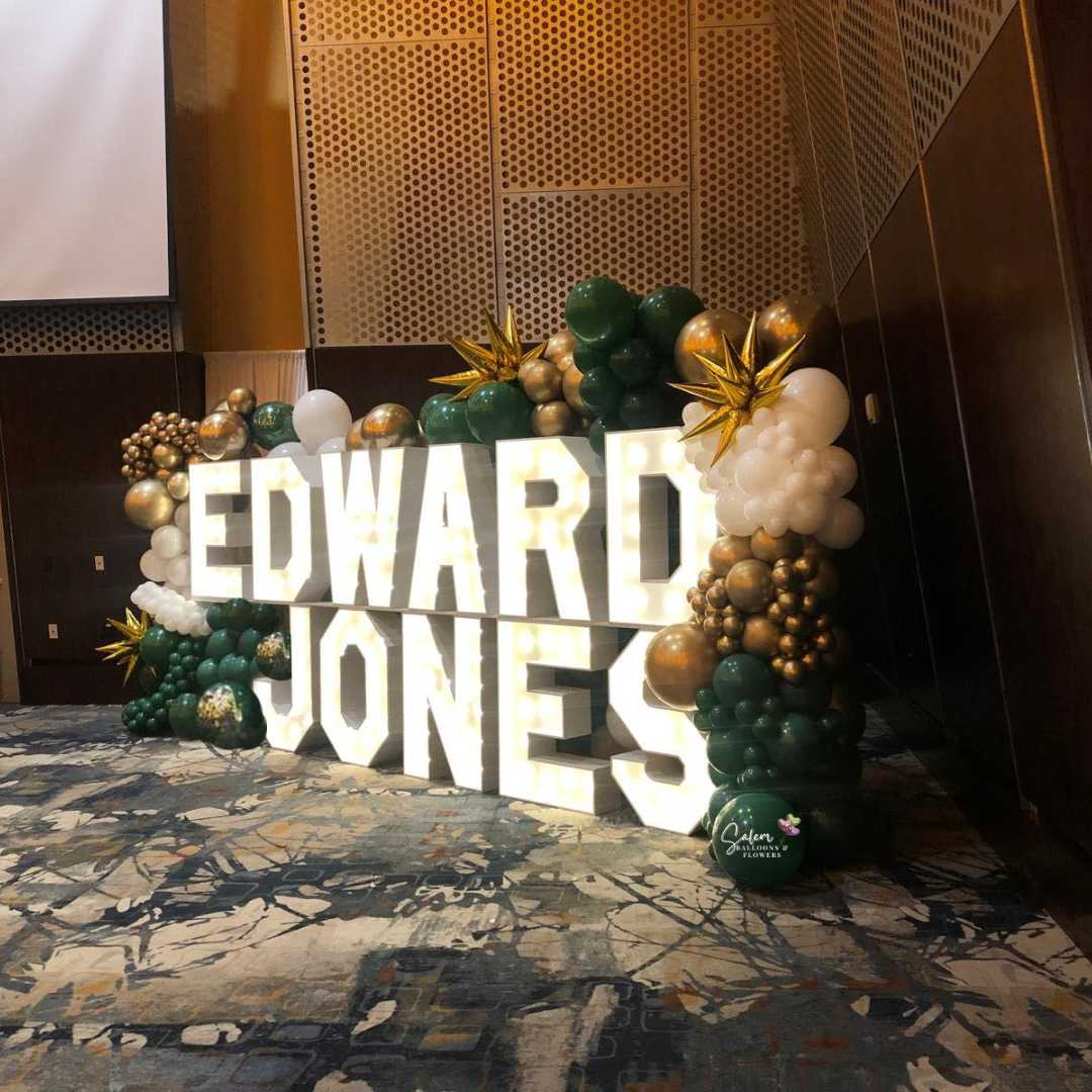 Light-Up marquee letters display spelling Edward Jones, nested in a 10 ft tall organic balloon wall in gold, green, and white. Salem Oregon Balloon Decor at the Salem Convention Center.