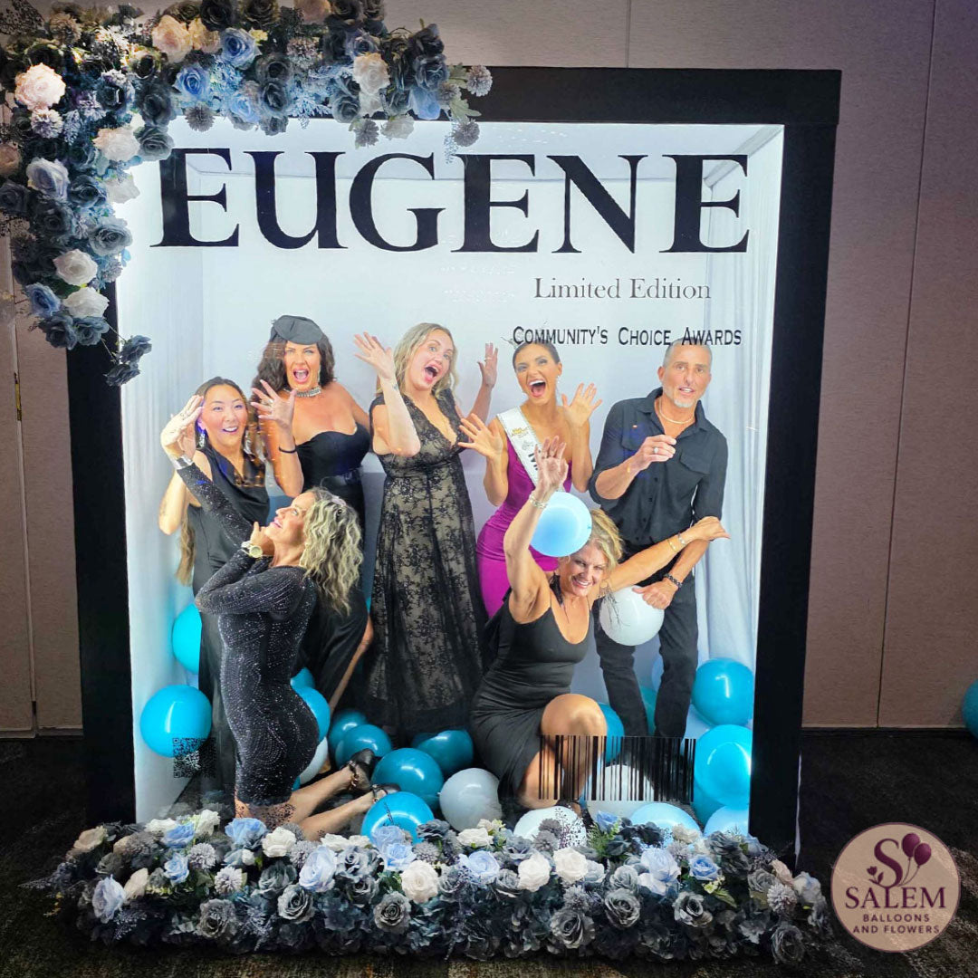 A group of 7 people posing together inside of a custom magazine photo booth featuring the text 'EUGENE Limited Edition COMMUNITY'S CHOICE AWARDS with blue and white balloons, floral decorations, and a 'Salem Saloon & Flowers' logo in the bottom right corner.