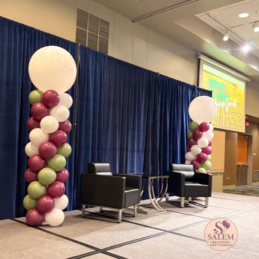 A corporate event decor setup in a conference room at the Salem Convention Center, featuring two tall balloon columns with white, burgundy, and sage green balloons, flanking a dark blue drape backdrop with two black armchairs and a small table between them, by Salem Balloons and Flowers.
