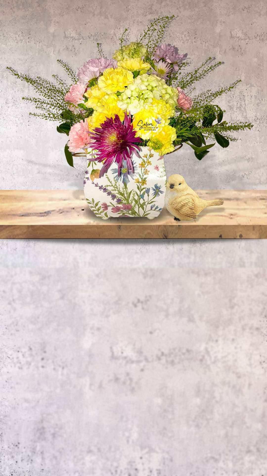 A Stunning flower arrangement in a pastel colors square vase with silvester flowers printed and a cute yellow bird standing next to it. Salem Oregon Flower delivery