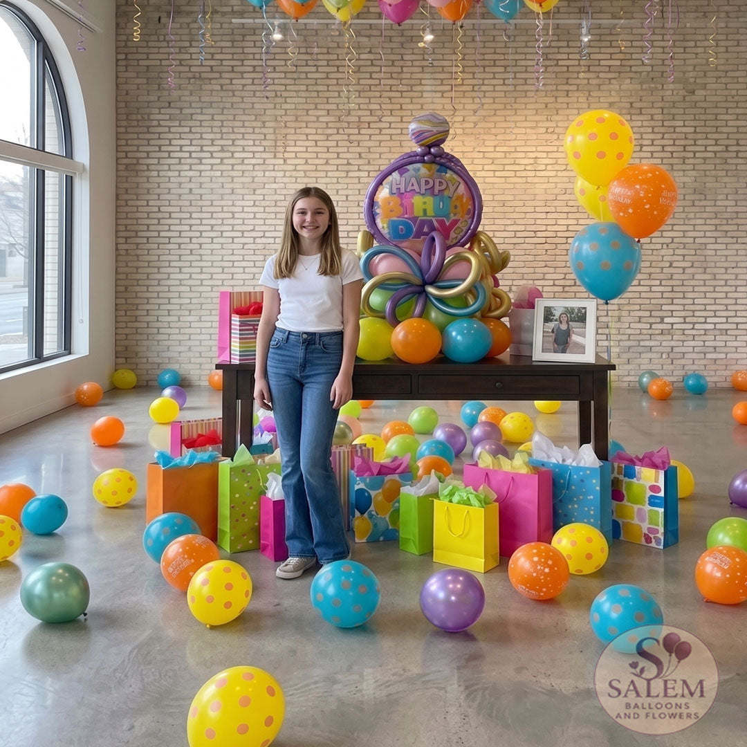 A young teenage girl in a white t-shirt and jeans stands next to a large, colorful 'Happy Birthday' balloon bouquet setup on a dark wood table in a bright room with scattered floor balloons and a full ceiling of helium balloons. Birthday balloon delivery in Salem Oregon by Salem Balloons and flowers.