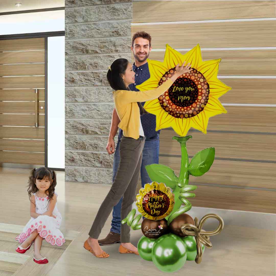 A mom standing next to her husband, daughter and an Extra tall Mother's day balloon bouquet. Featuring a Sunflower pick with a "Happy Mother's Day" message and decorated with curly balloons. Salem Oregon balloon delivery.
