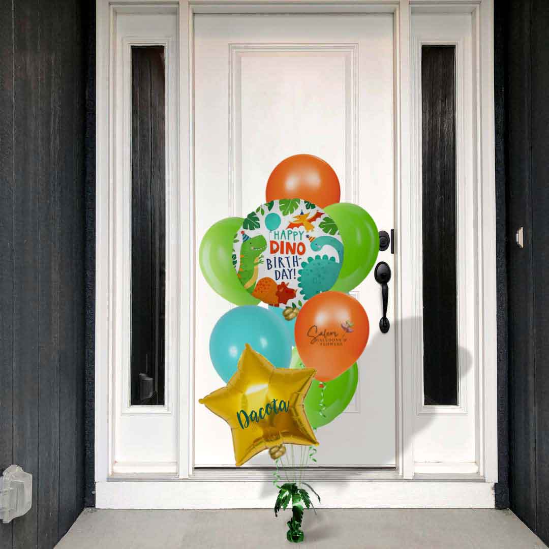 A white front door with a Dinosaur themed balloon bouquet. In a vibrant orange, green and caribbean blue palette. Featuring a "Happy Dino Birthday" message and a palm shaped balloon weight. Balloon delivery Salem Oregon and nearby cities.