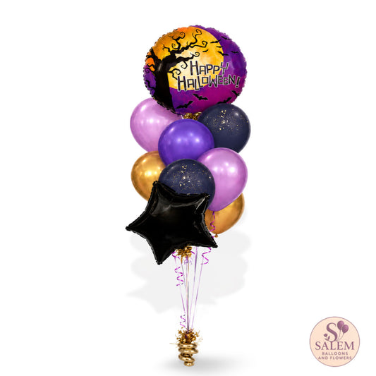 A balloon bouquet featuring a round 'Happy Halloween' foil balloon with a black tree silhouette over a purple and orange sky, surrounded by purple, lavender, navy, gold, and black balloons, tied together with a gold spool of ribbon on a white background.