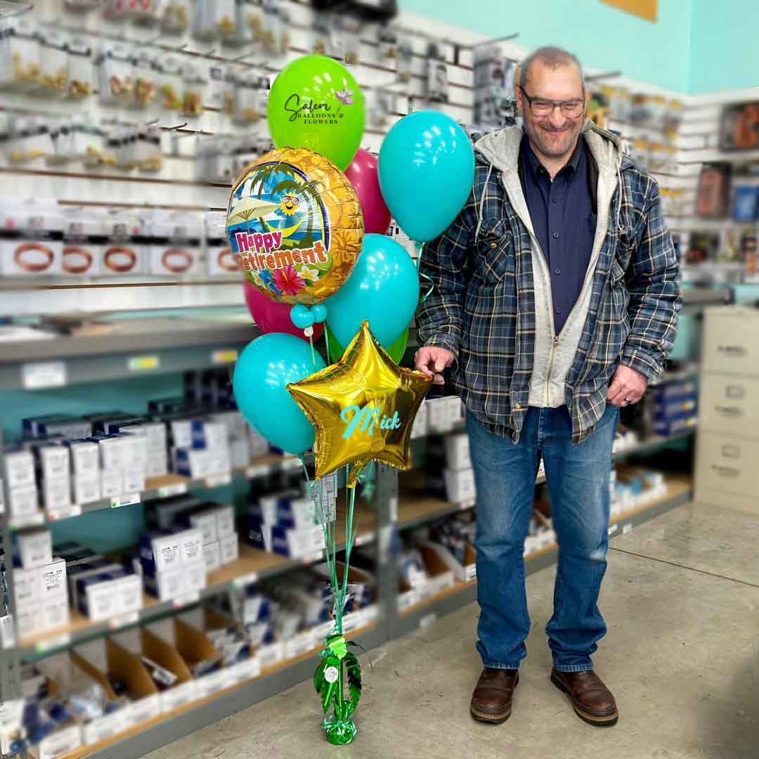 A smiling senior man standing next to a Hawaii-themed "Happy Retirement" Balloon Bouquet in colorful colors. Balloon weight decorated with a flower. Salem -Keizer Oregon balloon delivery