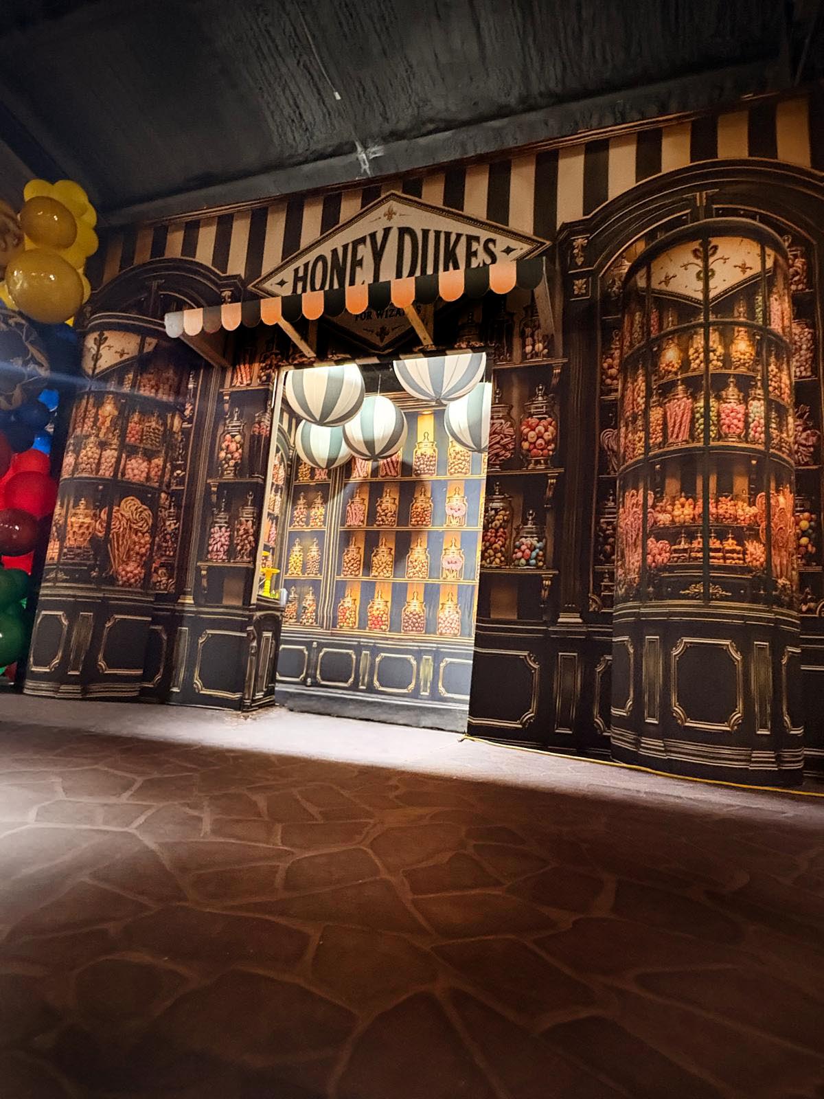 A decorative storefront-style backdrop with the sign 'HONEY DUKES' above striped walls, illuminated display panels, hanging lanterns, and a stone-tile floor.