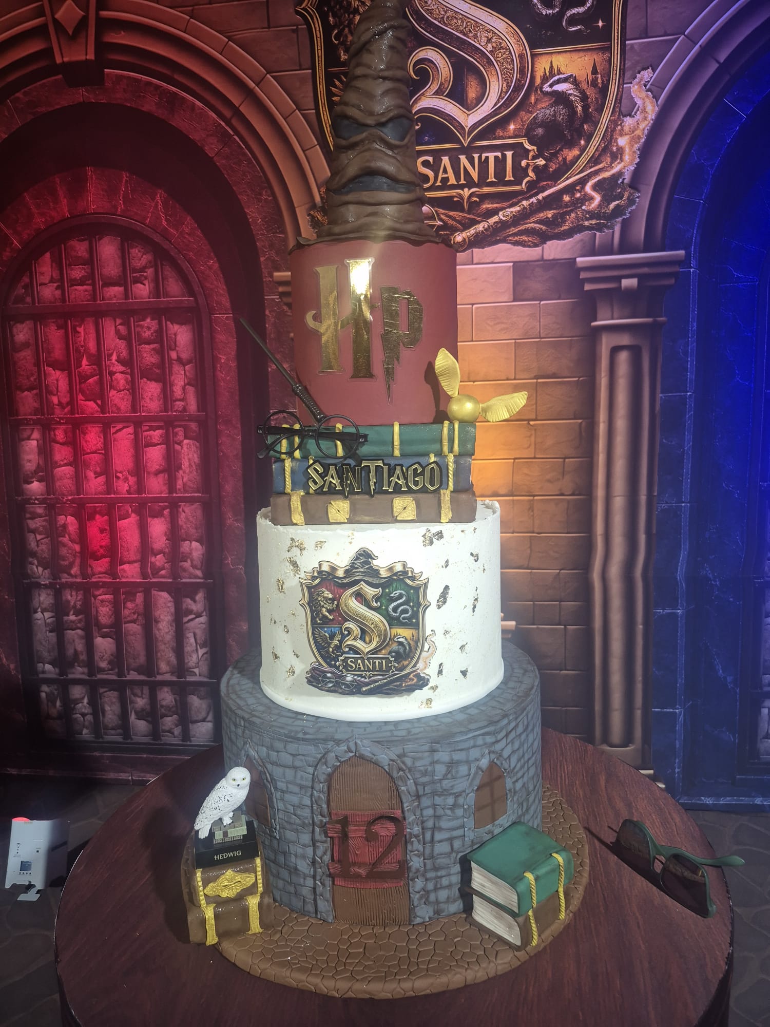 A highly detailed, multi-tiered wizard themed cake shaped like a stone tower, decorated with Slytherin and Gryffindor house crests, a golden snitch, and themed props, set against a dark, atmospheric studio backdrop resembling Hogwarts.