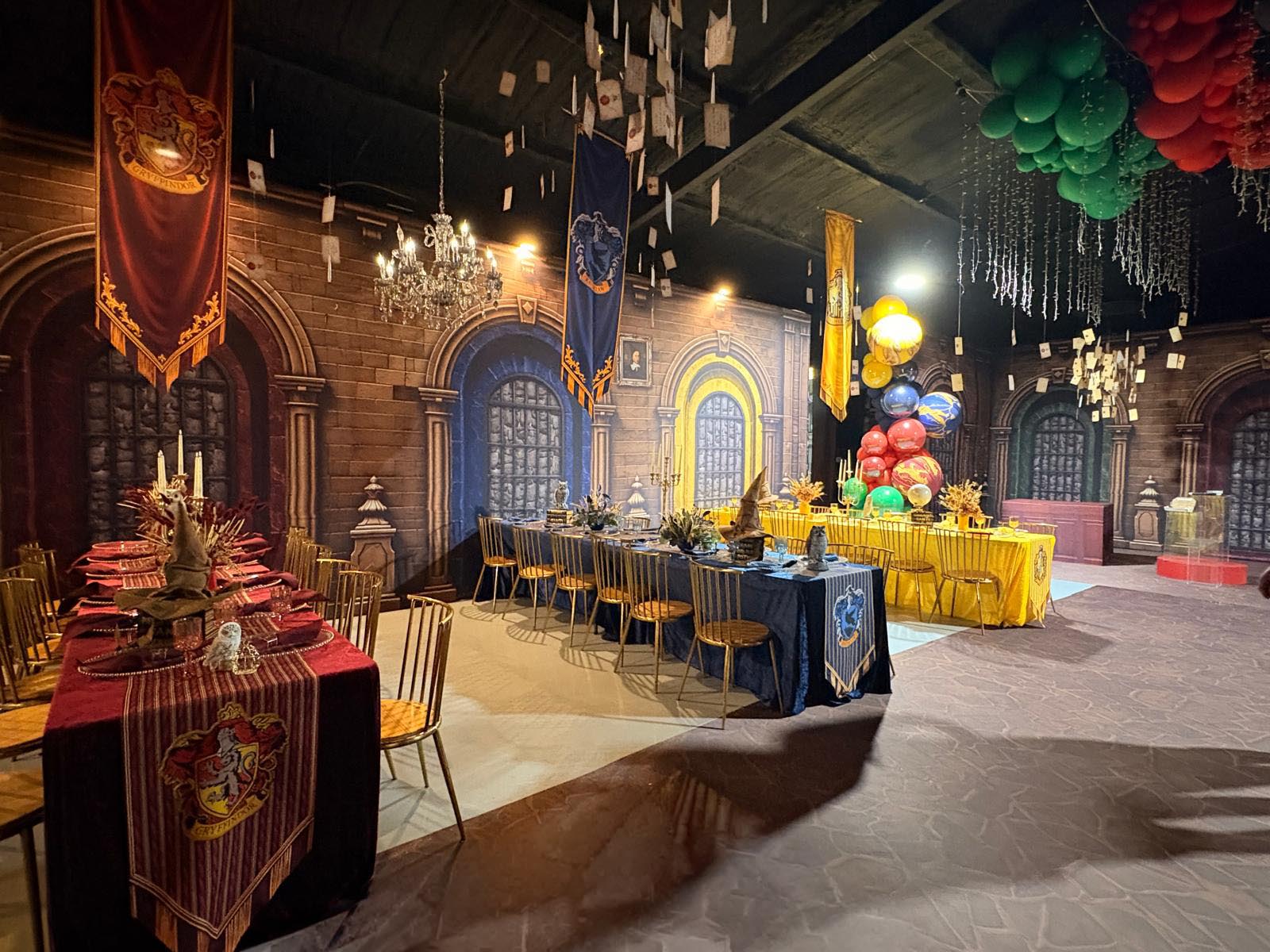 A themed event space decorated like a fantasy castle hall, with long tables set for dining, blue and red banners, hanging lanterns, and clusters of red, green, and yellow balloons overhead.