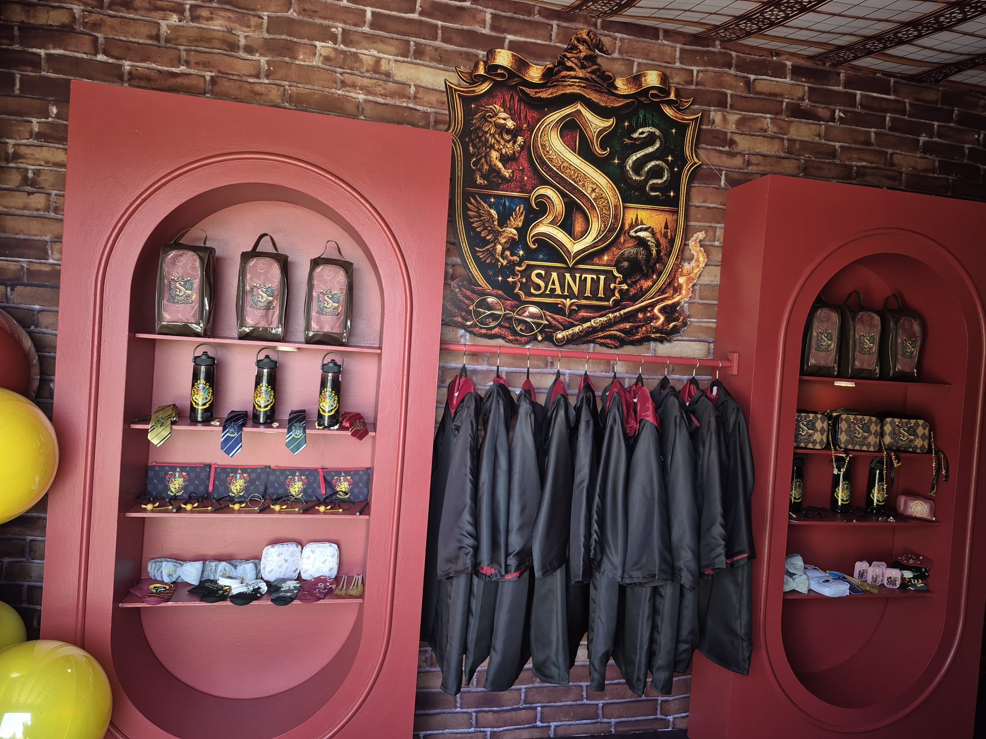 A retail display featuring a large central shield crest with the word 'SANTI' and a gold "S", flanked by two red arched shelving units holding decorative lanterns, small party favors accessories, and dark robes hanging on a rack against a brick wall.