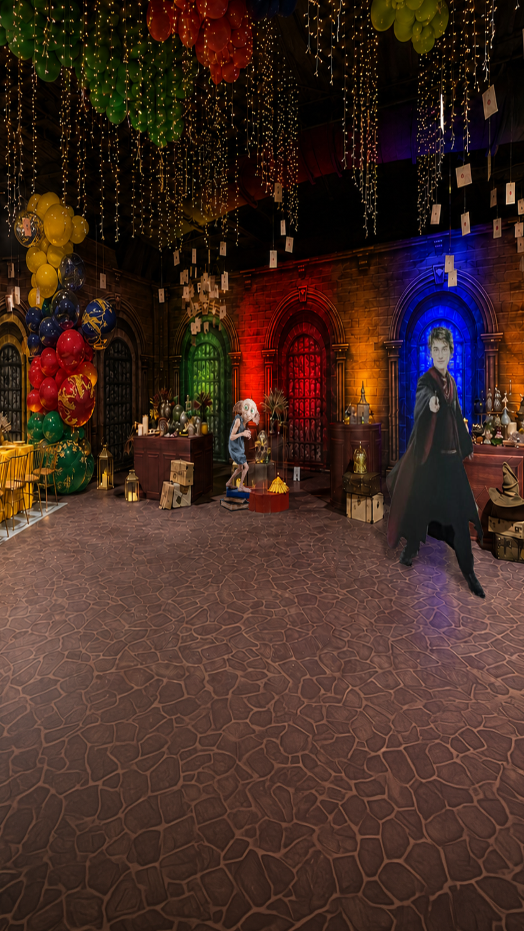 Immersive wizard-themed event environment with scenic architecture and custom décor with a gothic-themed party backdrop featuring a stone floor, arched windows with red and blue lighting, hanging fairy lights, balloons, and a character figure dressed in a black cape.