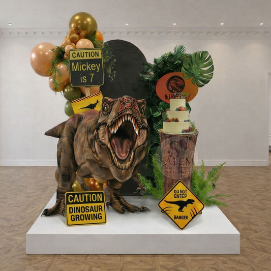 Dinosaur-Jurassic Park themed birthday balloon decoration, with a cake stand, greenery, balloon garlands, 2 panels, gian t-rex cutout, and signs in a decorated room. Balloon decor Oregon.
