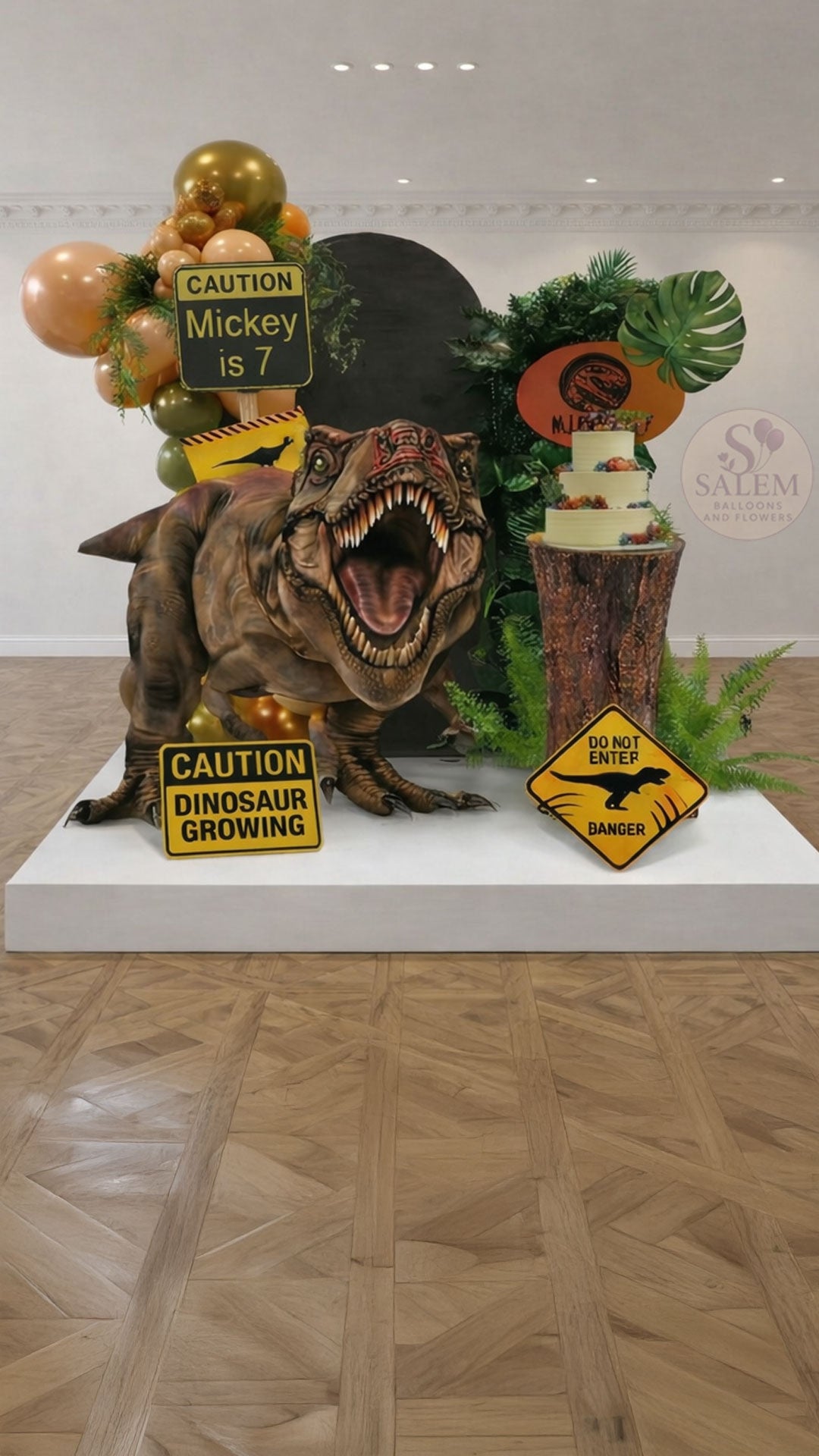 Dinosaur-Jurassic Park themed birthday balloon decoration, with a cake stand, greenery, balloon garlands, 2 panels, gian t-rex cutout, and signs in a decorated room. Balloon decor Oregon.