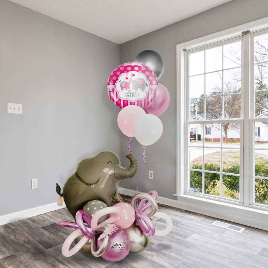 A Elephant themed balloon bouquet in a Pink and Gray color palette. Featuring a large elephant balloon holding a helium-filled balloon bouquet with a "It's a Girl" message, standing next to a window in a corner. Oregon Balloon delivery