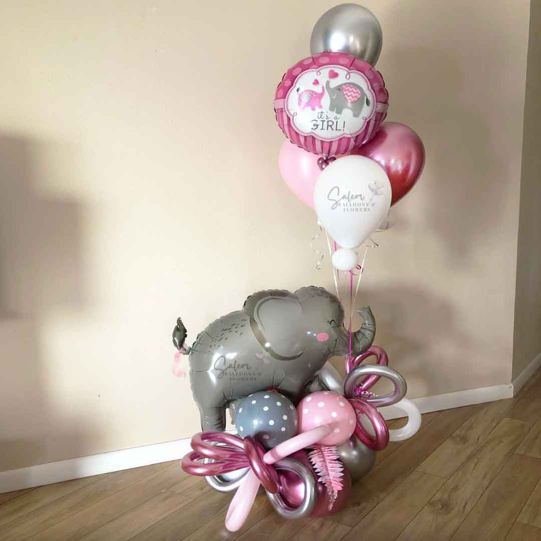 A Elephant themed balloon bouquet in a Pink and Gray color palette. Featuring a large elephant balloon holding a helium-filled balloon bouquet with a "It's a Girl" message, standing next to a window in a corner. Oregon Balloon delivery