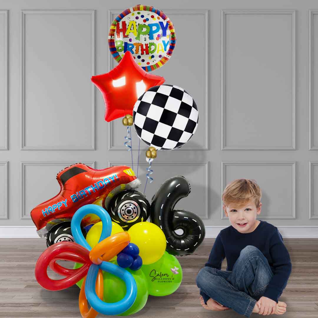 A boy sitting next to a balloon bouquet featuring a red monster truck balloon with a happy birthday message climbing a colorful balloon base with number balloon decorated as a road and a set of 3 helium balloons. Balloon delivery in Salem Oregon and nearby cities.