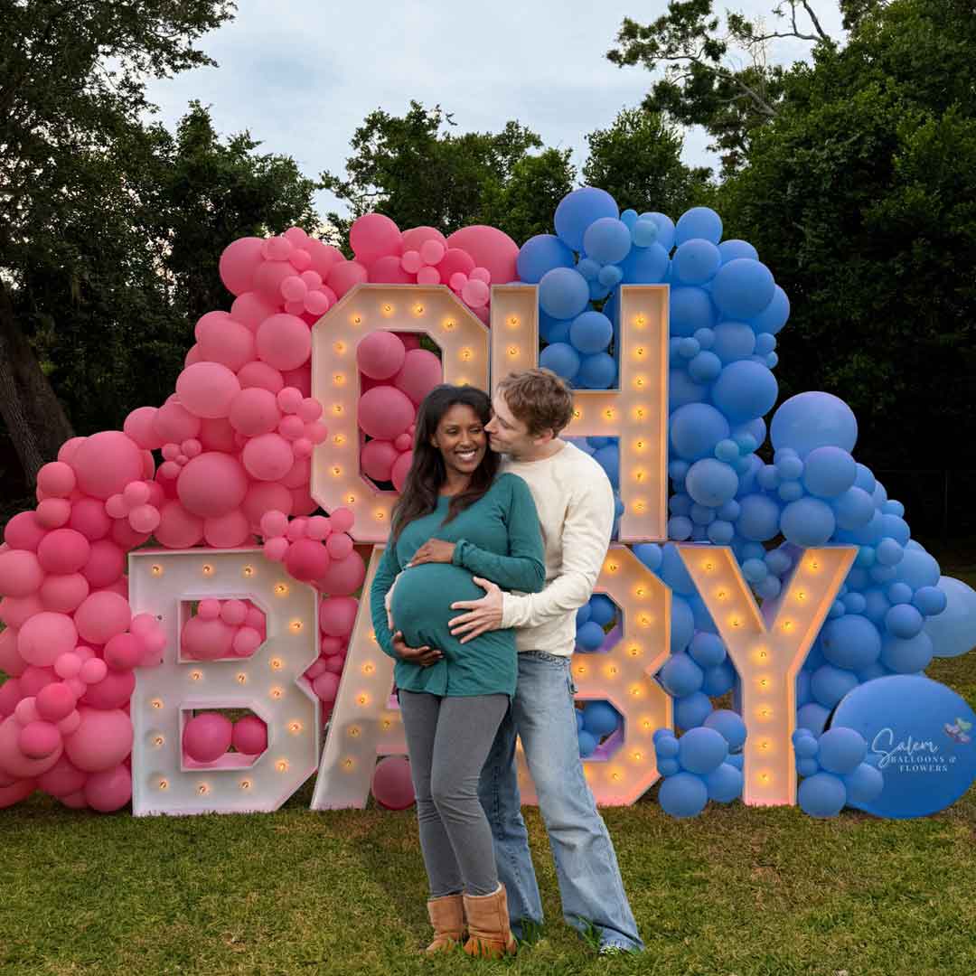 A couple standing in front of an "OH Baby" Gender Reveal Marquee Letter Display. 3ft Marquee letters nested into a pink and blue balloon wall. Balloon Decor and rentals in Oregon.