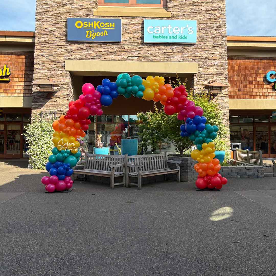 Colorful Organic balloon arch in front of a store with 'OshKosh B'gosh' and 'Carter's' signs at the Woodburn Outlets in Woodburn Oregon by Salem Balloons and flowers.