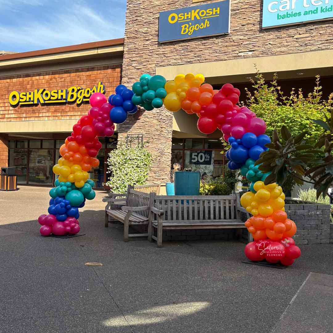 Colorful Organic balloon arch in front of a store with 'OshKosh B'gosh' and 'Carter's' signs at the Woodburn Outlets in Woodburn Oregon by Salem Balloons and flowers.