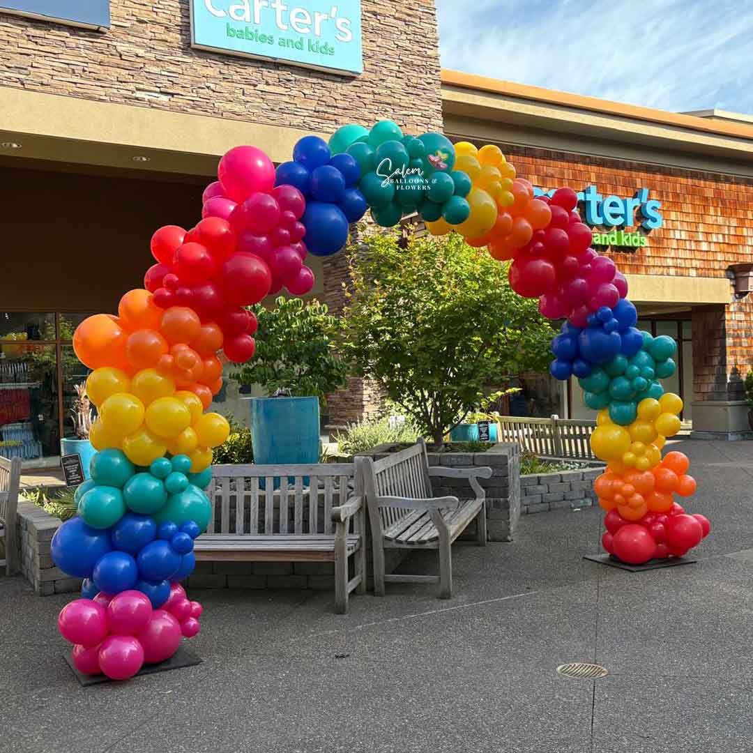 Colorful Organic balloon arch in front of a store with Carter's' signs at the Woodburn Outlets in Woodburn Oregon with a  Salem Balloons and flowers logo.