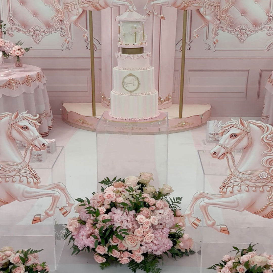 A pink and white carousel-themed party backdrop setup featuring a tiered cake on a platform, large white carousel horse graphics on both sides, and pink floral arrangements in the foreground.