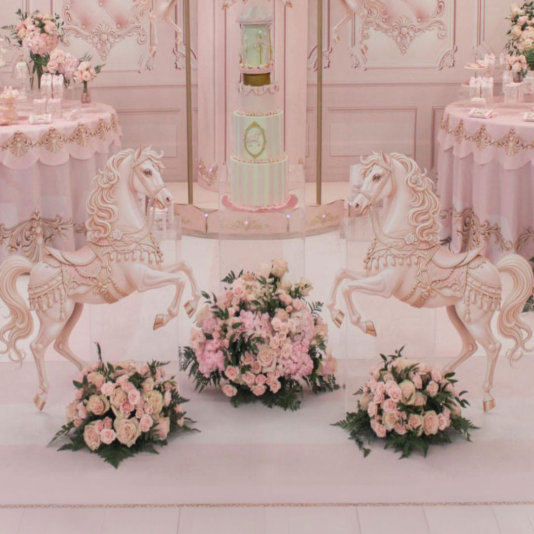 A pink baby shower party setup featuring two large pink decorative horse figures, a pastel pink tiered cake, and multiple floral arrangements on a light pink table runner.