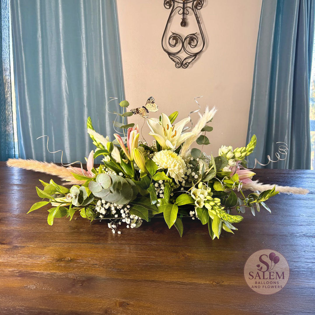 Floral arrangement with greenery, lily flowers, snapdragon flowers, curly twigs, and a butterfly with small pampas grass on a dining table, featuring the Salem Balloons and Flowers logo. Flower delivery in Salem Oregon and nearby cities.