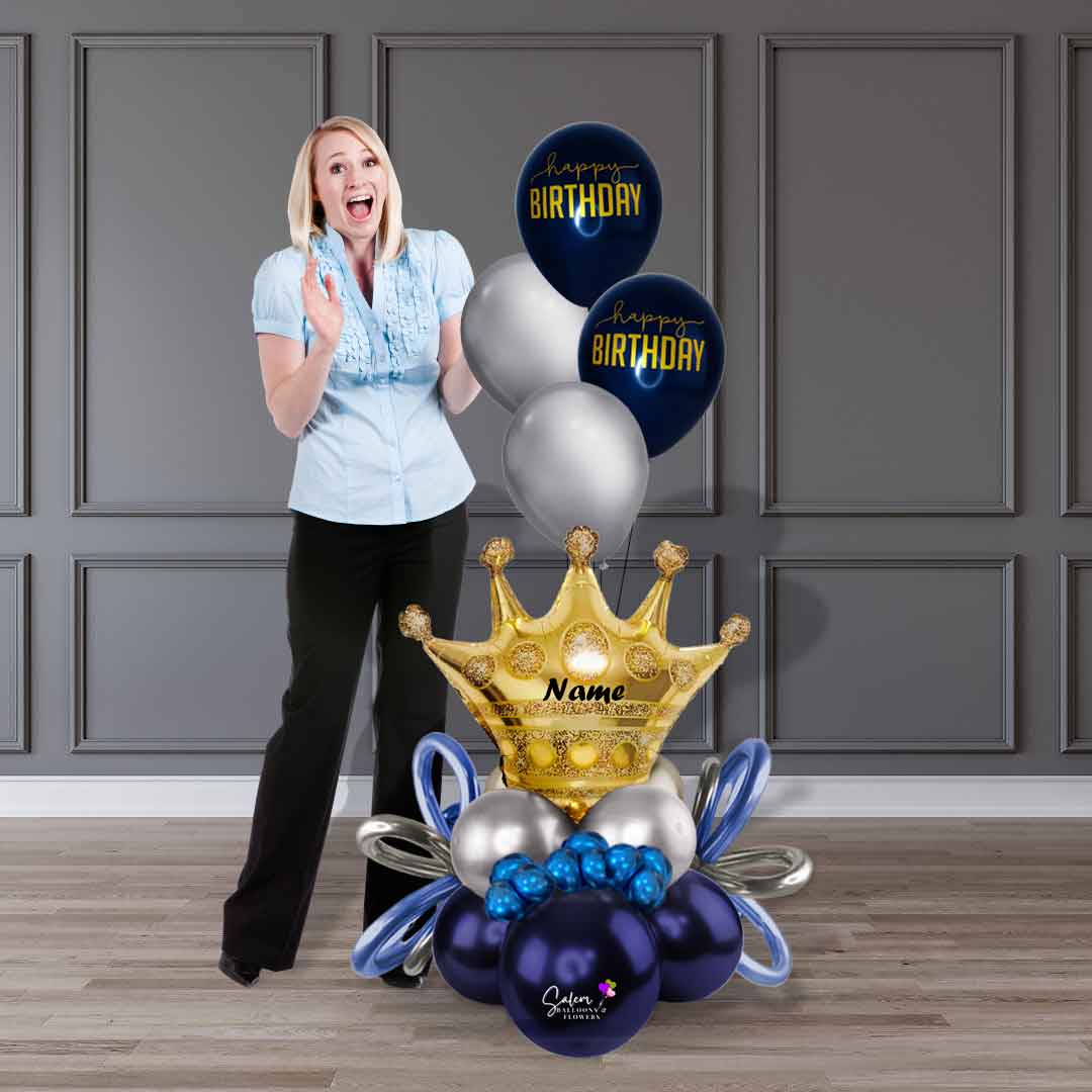 A happily surprised lady standing next to a birthday balloon bouquet with a large gold crown and helium balloons. Salem Oregon balloon delivery.