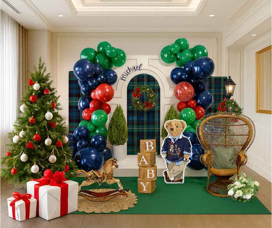 Decorative setup with balloon garlands in navy blue, red, and green, a Christmas tree, and teddy bear cutout, tall peacock chair and a rocking horse in a room. Baby shower balloon decor. Oregon.