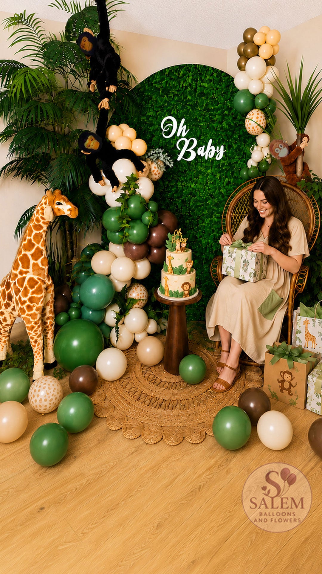 Expecting mother opening gifts in a safari-themed baby shower photo backdrop featuring a peacock chair, jungle balloon garland, hedge wall backdrop, giraffe prop, tropical greenery, and custom jungle cake by Salem Balloons and Flowers in Oregon.