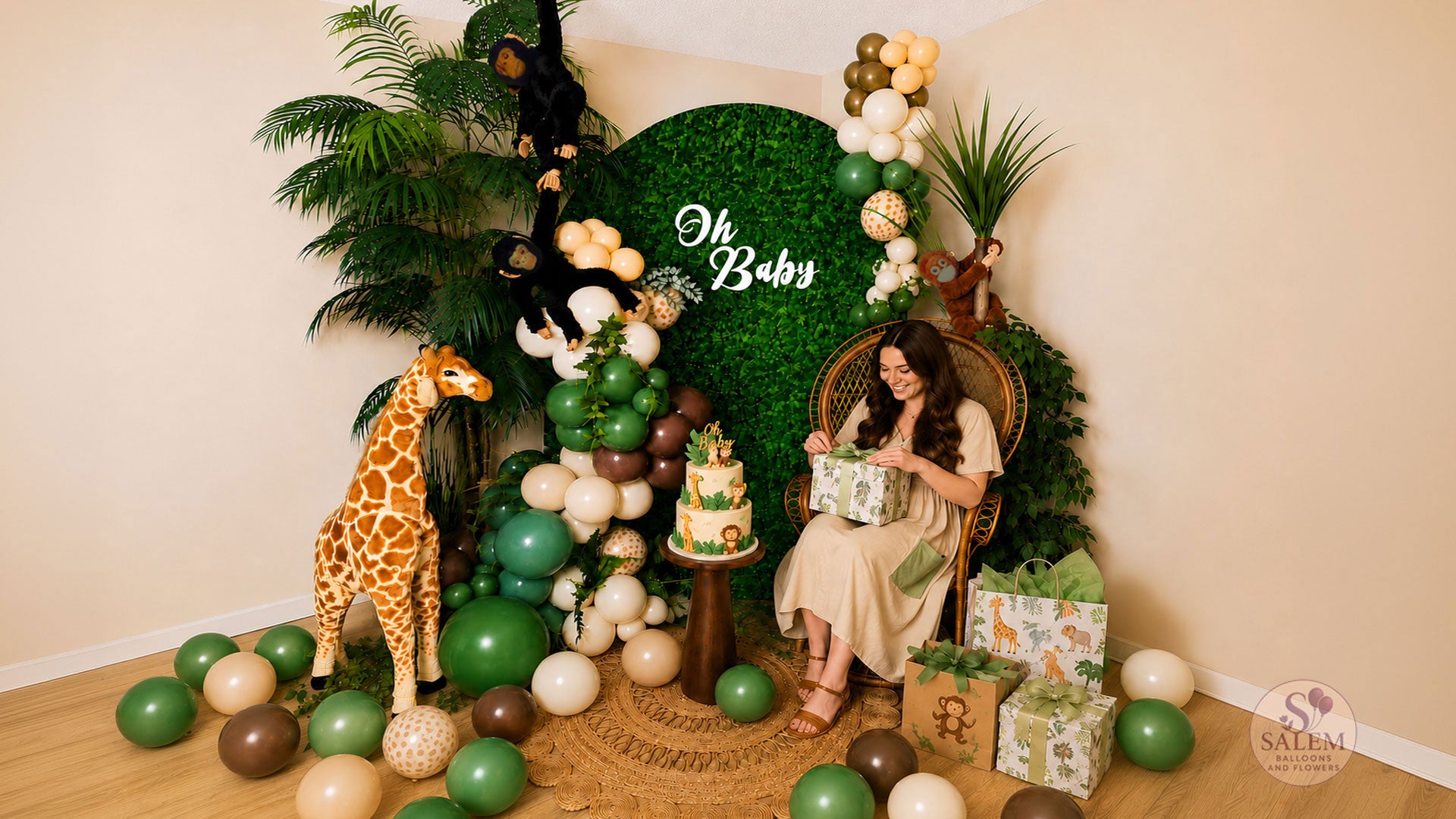 Expecting mother opening gifts in a safari-themed baby shower photo backdrop featuring a peacock chair, jungle balloon garland, hedge wall backdrop, giraffe prop, tropical greenery, and custom jungle cake by Salem Balloons and Flowers in Oregon.
