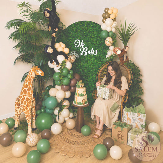 Expecting mother opening gifts in a safari-themed baby shower photo backdrop featuring a peacock chair, jungle balloon garland, hedge wall backdrop, giraffe prop, tropical greenery, and custom jungle cake by Salem Balloons and Flowers in Oregon.